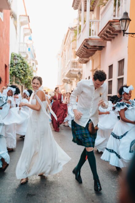La novia baila sonriente por una calle de balcones de madera, junto al novio con kilt y bailarinas de blanco