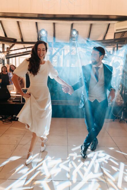 Los novios bailan de la mano en la pista entre haces de luz, ella con vestido midi de manga abullonada