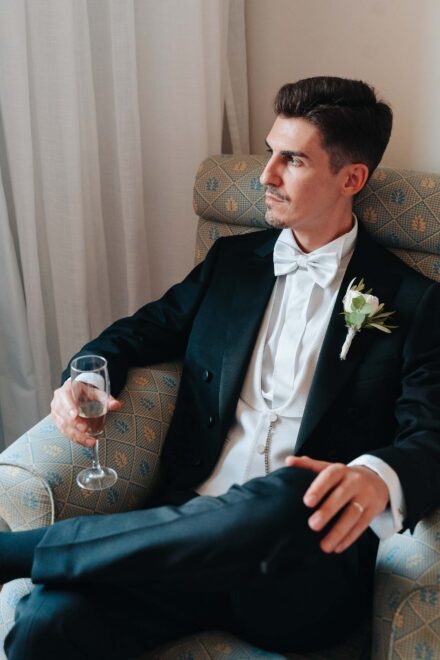 Elegant groom sitting in an armchair with a glass of champagne during wedding preparations in Seville