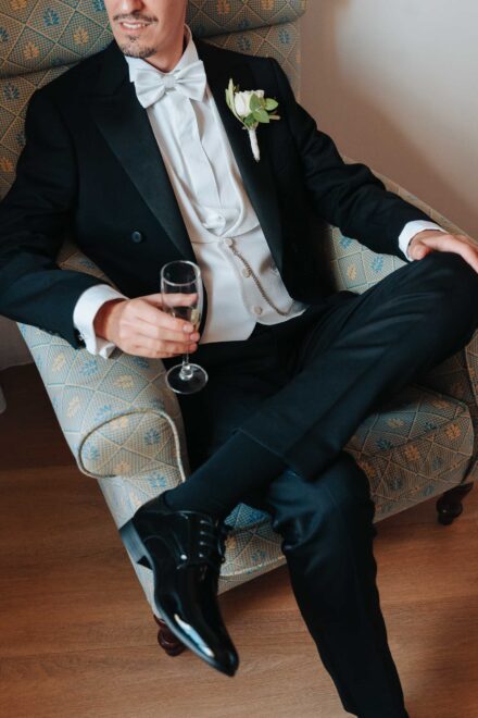 Groom in an elegant tuxedo sitting in an armchair with a glass of champagne during wedding preparations in Seville