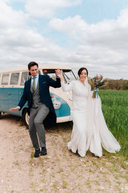 Los novios bailan de la mano delante de una furgoneta antigua azul y blanca, junto a un campo verde