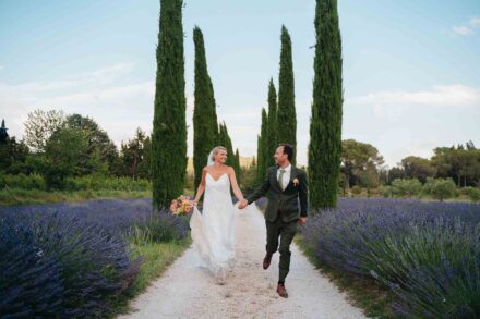 Los novios corren de la mano por un camino de grava entre cipreses y campos de lavanda