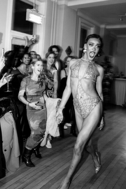 Una artista con body de pedrería desfila sacando la lengua ante los invitados, que gritan detrás, en blanco y negro