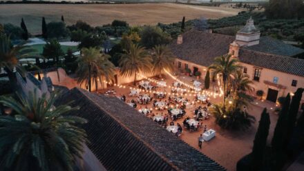 Vista aérea del banquete en el patio de una hacienda al anochecer, con palmeras y guirnaldas de luces