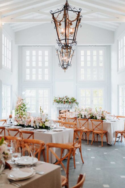 Salón blanco de ventanales altos con faroles de forja colgados y mesas con flores y velas