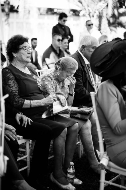 Una invitada mayor se seca las lágrimas con un pañuelo en la ceremonia, junto a otra señora, en blanco y negro