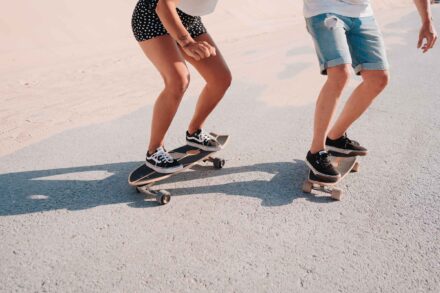 Las piernas de la pareja, con zapatillas negras, montando en monopatín por una carretera junto a la arena