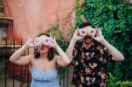 La pareja se tapa los ojos con dos donuts rosas cada uno, frente a una pared cubierta de hiedra