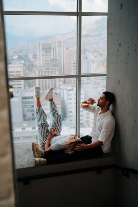 La novia apoya las piernas en el ventanal con una porción de pizza mientras el novio bebe, ante una ciudad entre montañas