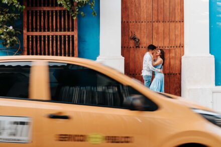 La pareja se abraza ante un portón de madera en una fachada azul mientras pasa un taxi amarillo movido