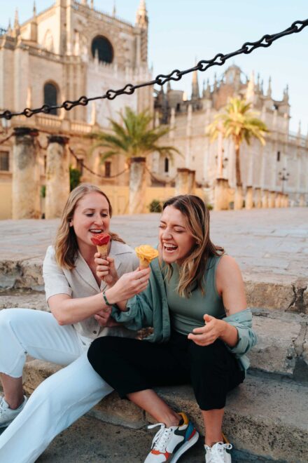 Dos chicas se ríen comiendo helado sentadas en unas gradas, frente a la catedral de Sevilla