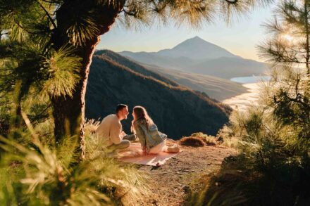 La pareja, sentada en una manta entre pinos, a punto de besarse frente a un volcán y un mar de nubes