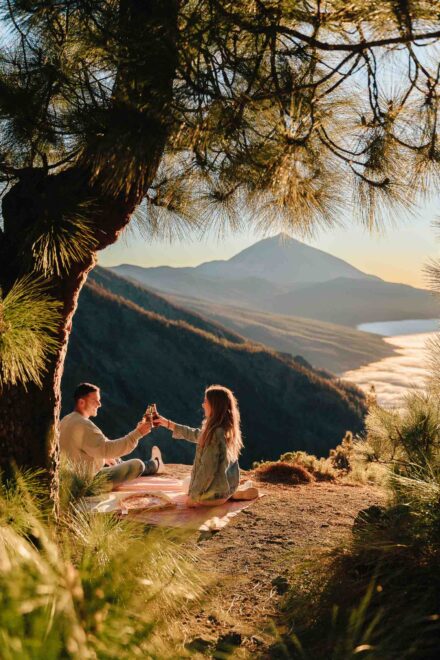 La pareja brinda con botellines sobre una manta bajo un pino, con el Teide y un mar de nubes al fondo