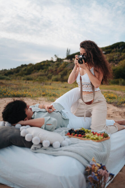 La novia fotografía con una cámara analógica al novio, tumbado en un colchón de pícnic junto a una bandeja de fruta