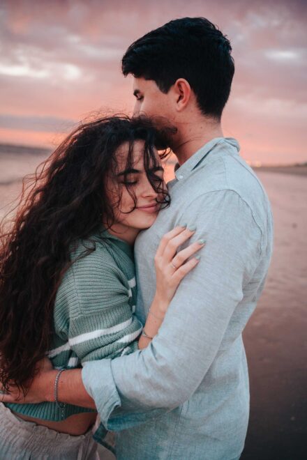 La pareja se abraza en la playa bajo un cielo rosa; ella apoya la cabeza en su pecho con los ojos cerrados