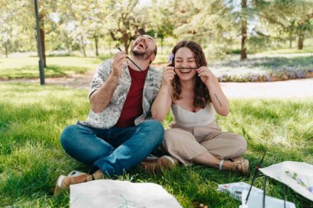 La novia se pone un pincel de bigote y el novio se ríe, sentados en el césped junto a sus láminas pintadas