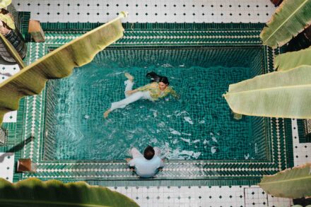 Vista cenital de la novia nadando vestida en una piscina de azulejos verdes mientras el novio la mira