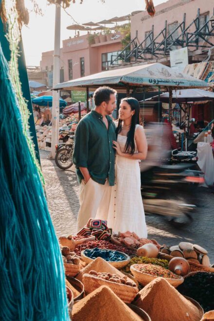 La pareja se mira muy junta en un mercado, tras unos cestos de especias, mientras pasa una moto
