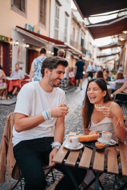 Los novios se ríen, cada uno con un pastel de nata, en la terraza de un café de una calle empedrada