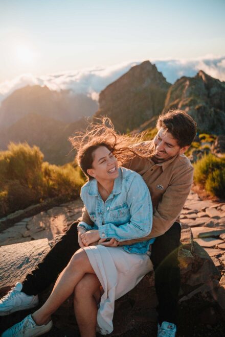 La novia ríe con el pelo al viento, sentada en el regazo del novio en una cumbre sobre las nubes