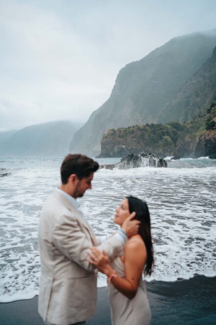 El novio sujeta la cara de la novia en la orilla, desenfocados ante las olas y la montaña con niebla