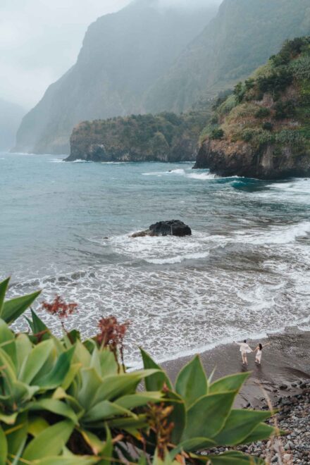 La pareja camina de la mano hacia el mar en una playa de arena oscura entre acantilados verdes, vista desde lo alto