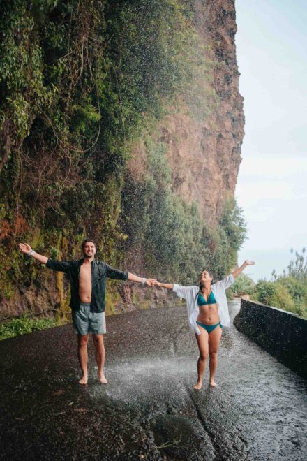 Los novios, en bañador y de la mano, abren los brazos bajo una cascada que cae sobre la carretera