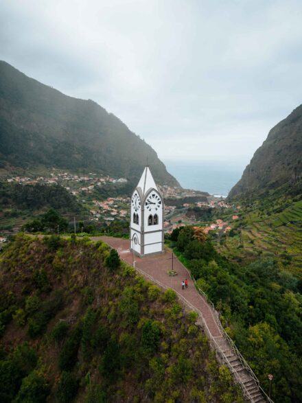 Vista aérea de la pareja paseando junto a un campanario blanco sobre un valle que baja hasta el mar