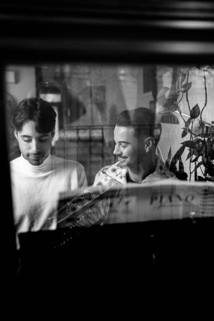 Los novios leen una partitura al piano, vistos a través de una ventana, en blanco y negro