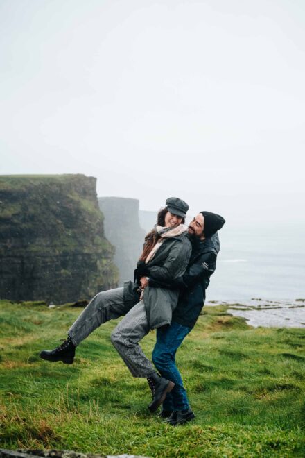 El novio levanta a la novia por la cintura, ella con gorra y bufanda, en unos acantilados verdes con niebla