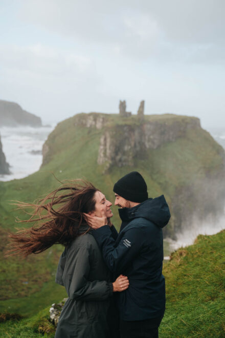 Pareja abrazada en su preboda en un acantilado junto al mar, ella con el pelo al viento, con un castillo en ruinas detrás