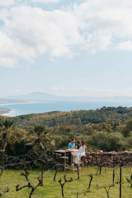 Los novios en una mesa alta junto a un muro de piedra, entre viñas y palmitos, con el mar y la costa al fondo