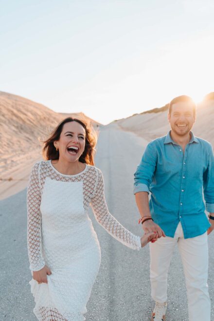 Los novios caminan de la mano y ríen por una carretera entre dunas, con el sol bajo a contraluz