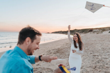 La novia sostiene en alto una cometa blanca y ríe en la playa al atardecer, con el novio desenfocado delante