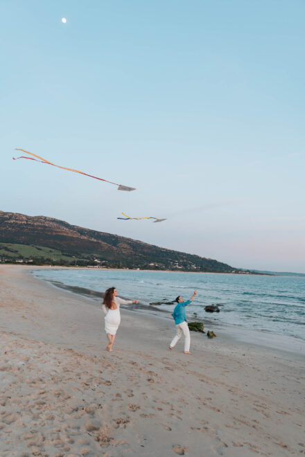 Los novios vuelan dos cometas de colas de colores en una playa larga al atardecer, con la luna en el cielo