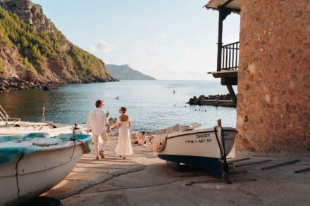 Los novios, de blanco y de espaldas, bajan por una rampa entre barcas varadas hacia una cala con bañistas