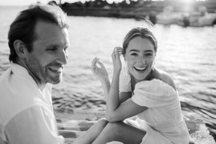 La novia ríe mirando a cámara sentada junto al mar, con el novio sonriendo a su lado, en blanco y negro