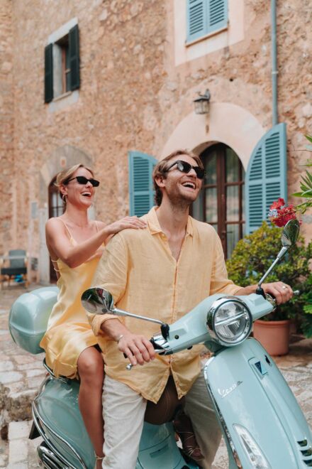 Los novios se ríen con gafas de sol subidos a la moto frente a una casa de piedra con contraventanas azules