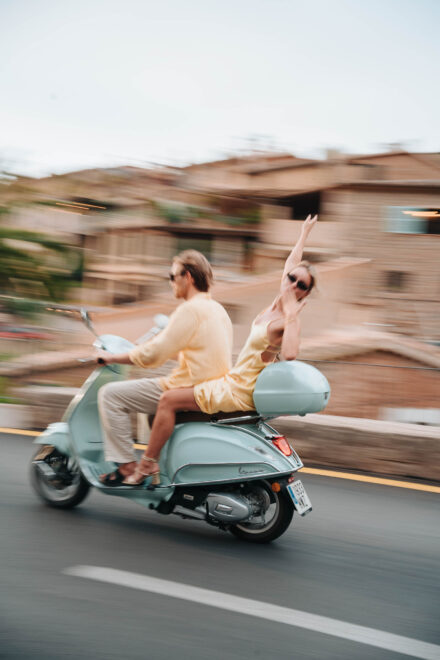 Los novios pasan en una moto azul claro con el fondo barrido por la velocidad, ella con un brazo en alto