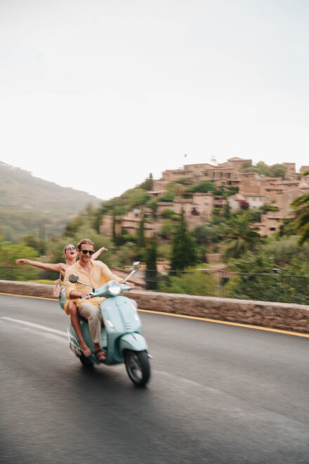 Los novios van en moto por la carretera, ella con los brazos abiertos y un pueblo de piedra en la colina