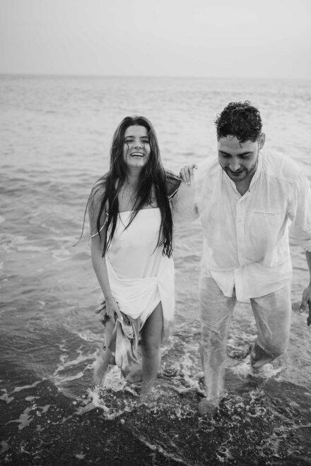 Los novios salen del agua empapados y riendo, ella con el vestido blanco recogido, en blanco y negro