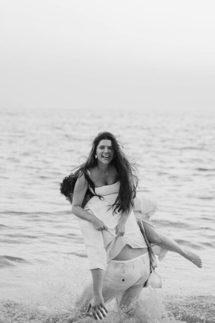 El novio carga con la novia dentro del mar mientras ella ríe con el pelo al viento, en blanco y negro