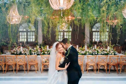Los novios abrazados en la pista, frente a la mesa del banquete, bajo lámparas de cristal y plantas colgantes