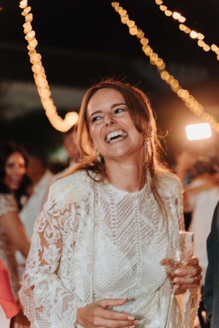 La novia, con vestido de encaje de manga larga, se ríe con una copa en la mano bajo guirnaldas de luces