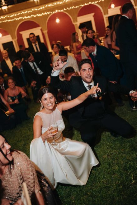 De noche, la novia, sentada en el césped con una copa, y el novio, de rodillas, levantan los brazos entre los invitados