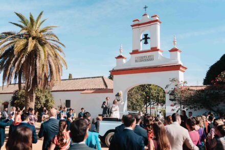Los novios saludan de pie en un coche antiguo, ella con el ramo en alto, en la entrada del Cortijo El Chamorro