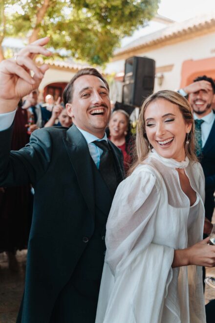Los novios ríen a carcajadas rodeados de invitados en un patio; el novio levanta una mano