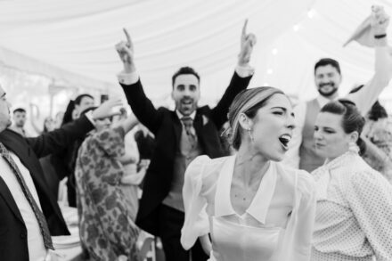 La novia canta a pleno pulmón entre invitados con los brazos en alto bajo una carpa, en blanco y negro