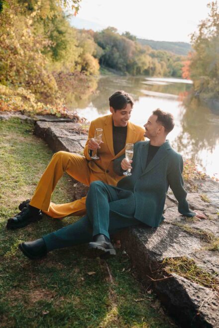 Los novios, con trajes mostaza y verde, brindan con champán sentados en unas rocas junto a un río en otoño