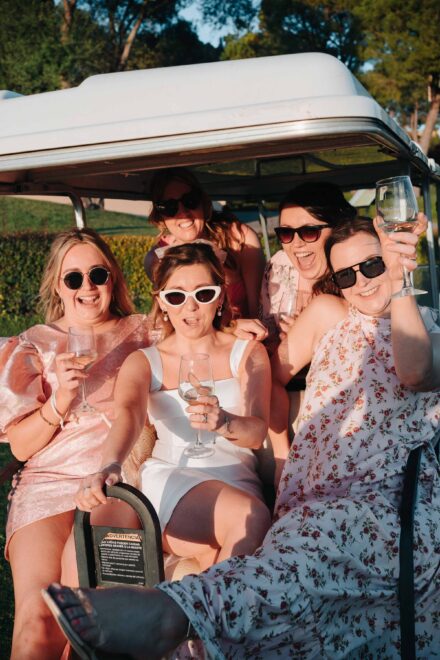 La novia, con gafas de sol blancas, posa con cuatro amigas y sus copas de vino, apretadas en un carrito de golf al sol